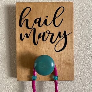 May include: Wooden sign with the words "hail Mary" in black script. A teal knob is attached to the sign, holding a pink beaded necklace with blue accents. The sign is mounted on a light-colored wall.