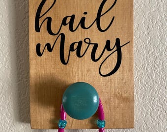 May include: Wooden sign with the words "hail Mary" in black script. A teal knob is attached to the sign, holding a pink beaded necklace with blue accents. The sign is mounted on a light-colored wall.