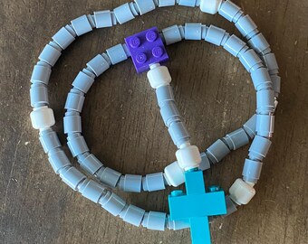 May include: A rosary made of gray and white toy blocks, with a purple block at the center and a blue cross at the bottom. The blocks are connected to form a loop.