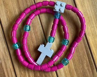 May include: A pink and blue Lego necklace with a white cross charm. The necklace is made of small plastic beads.