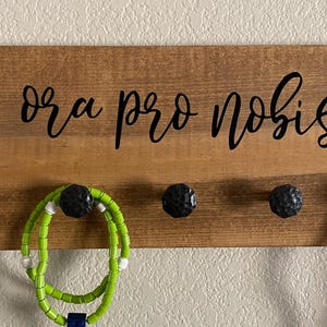 May include: Wooden wall-mounted jewelry organizer with the phrase "ora pro nobis" in black script. The organizer has four black knobs for hanging jewelry. Various necklaces and bracelets are displayed on the hooks, including beaded and colorful pieces.