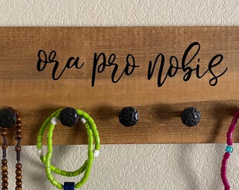 May include: Wooden wall-mounted jewelry organizer with the phrase "ora pro nobis" in black script. The organizer has four black knobs for hanging jewelry. Various necklaces and bracelets are displayed on the hooks, including beaded and colorful pieces.