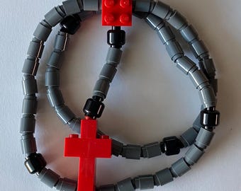 May include: A handmade rosary made from gray and black Lego bricks, with a red Lego cross pendant. The rosary features a series of gray cylindrical beads connected by black Lego pieces, and a red Lego square at the top.