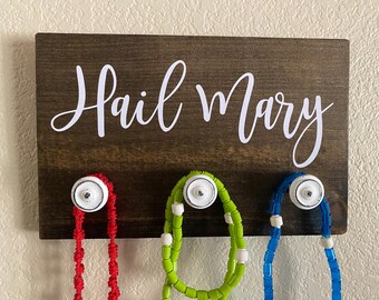 May include: A dark stained wooden sign with the words "Hail Mary" in white script. Below the sign are three white knobs with red, green, and blue beaded rosaries hanging from them.