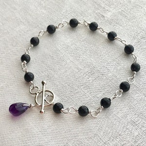 May include: A silver chain bracelet with black beads and a purple amethyst pendant. The bracelet has a toggle clasp.