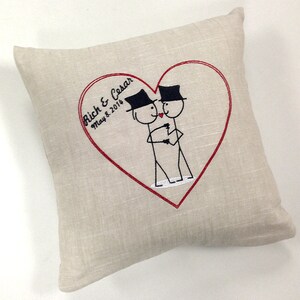 May include: A beige pillow with a red heart embroidered on it. Inside the heart are two stick figures wearing top hats, holding hands, and kissing. The text "Rich & Cesar May 8, 2014" is embroidered above the figures.