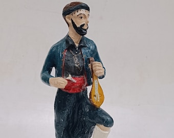 Pode incluir: Uma figura de um homem vestindo uma roupa azul e vermelha e um chapéu, segurando um instrumento musical. A figura está em pé sobre uma base marrom.