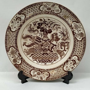May include: A white ceramic plate with a brown floral design. The plate features a central image of a vase with flowers and a dragon. The plate has a decorative border with a repeating pattern.