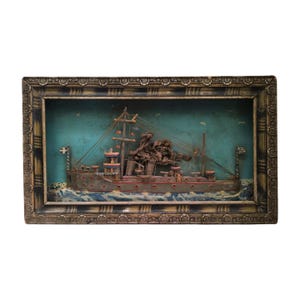 Könnte beinhalten: Ein gerahmtes Diorama eines Holzschiffs vor blauem und grünem Hintergrund. Das Schiff ist aus Holz und hat einen braunen Rumpf, weiße Segel und rote und weiße Akzente. Das Diorama befindet sich in einem Holzrahmen mit goldener Verzierung.