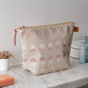 May include: A beige zippered pouch with a pink and white geometric pattern. The pouch has a pink zipper pull and a brown leather strap.