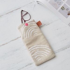 May include: A beige linen eyeglasses case with a white abstract design and a pink button closure. The case is lying on a white wooden surface.