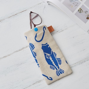 May include: A beige canvas glasses case with a blue fish and hook design. The case has a blue button closure and a brown leather tag.