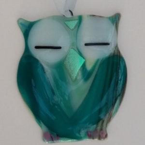 May include: A teal blue glass owl ornament with iridescent green accents and white eyes. The owl has a whimsical, sleepy expression.