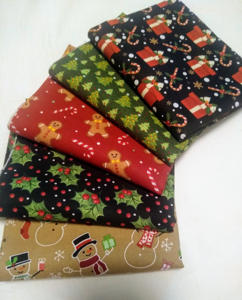 May include: A stack of five Christmas-themed fabric squares. The top square is black with red and white candy canes, presents, and snowflakes. The second square is green with white Christmas trees. The third square is red with white gingerbread men and candy canes. The fourth square is black with green holly leaves and red berries. The bottom square is brown with white snowmen and snowflakes.