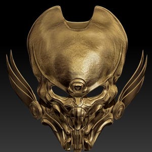 Puede incluir: Una máscara dorada, de diseño intrincado, con apariencia de calavera. La máscara presenta líneas detalladas, aberturas para los ojos y estructuras en forma de alas en los laterales. Un pequeño emblema de calavera está centrado sobre las aberturas de los ojos. La máscara tiene un acabado metálico.