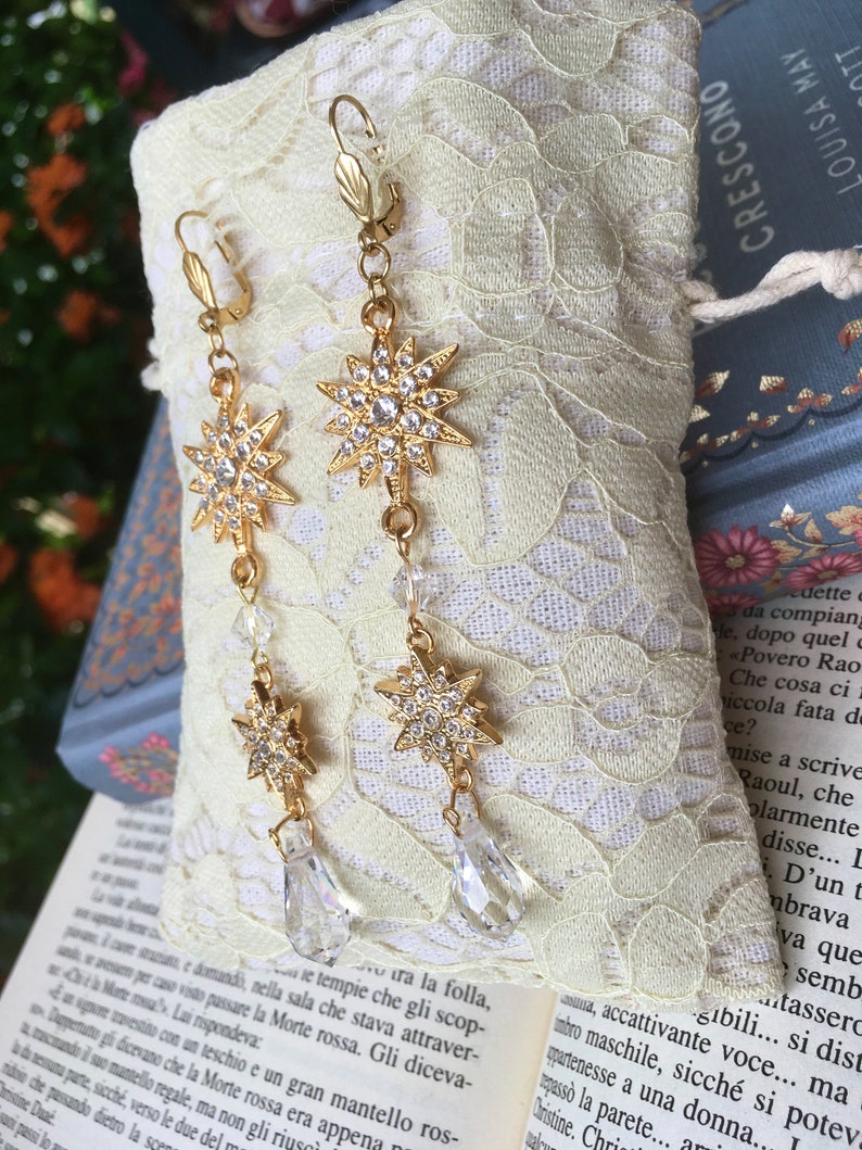 K&ouml;nnte beinhalten: Ein Paar goldene Ohrringe mit einem Sternendesign und klaren Kristall-Akzenten. Die Ohrringe h&auml;ngen an einer zarten Kette mit einem tropfenf&ouml;rmigen Kristall am unteren Ende.