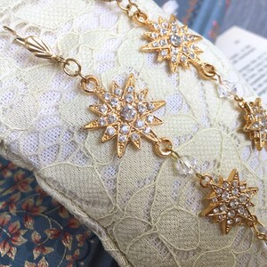 K&ouml;nnte beinhalten: Eine goldene Kettenkette mit mehreren sternf&ouml;rmigen Anh&auml;ngern, die mit klaren Strasssteinen verziert sind. Die Kette ist mit klaren Kristallperlen durchsetzt.