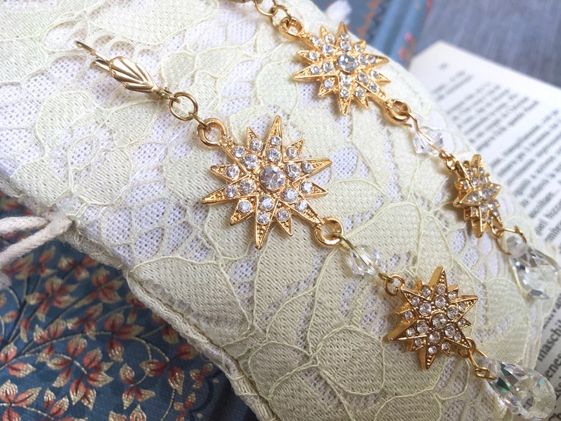 K&ouml;nnte beinhalten: Eine goldene Kettenkette mit mehreren sternf&ouml;rmigen Anh&auml;ngern, die mit klaren Strasssteinen verziert sind. Die Kette ist mit klaren Kristallperlen durchsetzt.