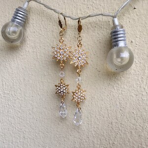 K&ouml;nnte beinhalten: Goldfarbene Ohrringe mit einem baumelnden Sternendesign und Kristall-Akzenten. Die Sterne sind mit klaren Strasssteinen verziert und die Ohrringe haben einen tropfenf&ouml;rmigen Kristall am unteren Ende.