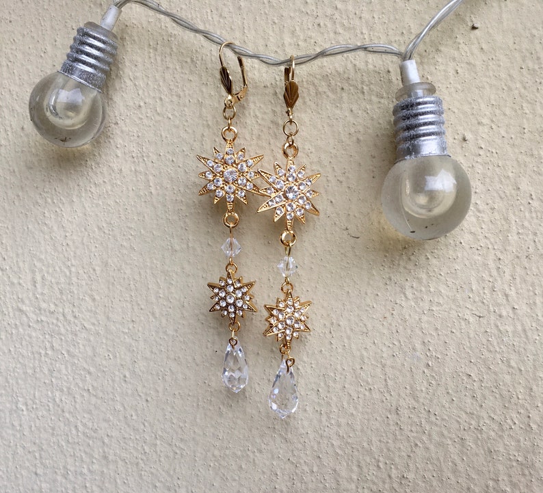 K&ouml;nnte beinhalten: Goldfarbene Ohrringe mit einem baumelnden Sternendesign und Kristall-Akzenten. Die Sterne sind mit klaren Strasssteinen verziert und die Ohrringe haben einen tropfenf&ouml;rmigen Kristall am unteren Ende.