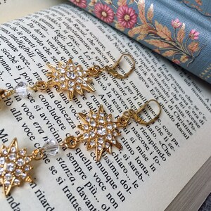 K&ouml;nnte beinhalten: Goldene sternf&ouml;rmige Ohrringe mit klaren Kristall-Akzenten. Die Ohrringe h&auml;ngen an einem Buch mit einem floralen Design.