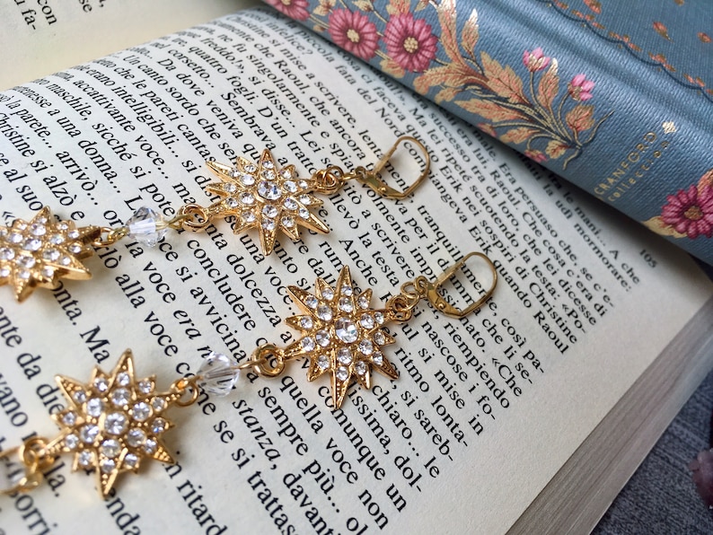 K&ouml;nnte beinhalten: Goldene sternf&ouml;rmige Ohrringe mit klaren Kristall-Akzenten. Die Ohrringe h&auml;ngen an einem Buch mit einem floralen Design.