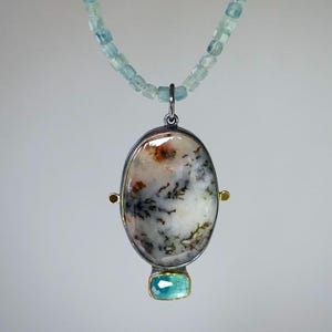Puede incluir: Un collar con una hilera de pequeñas cuentas azul claro facetadas. El colgante es una piedra ovalada con inclusiones dendríticas blancas, marrones y negras, engastada en un marco de metal oscuro con detalles dorados y una pequeña piedra azul rectangular.