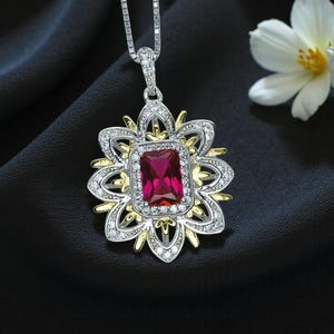 May include: A silver necklace with a flower-shaped pendant. The pendant features a rectangular, ruby-colored stone at its center, surrounded by a gold and silver floral design. The necklace is displayed on a black surface, with a white flower in the background.