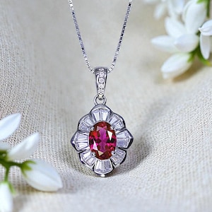 May include: A silver necklace with a pendant featuring a large oval ruby gemstone surrounded by smaller, rectangular diamonds. The pendant is suspended from a delicate chain. White flowers are in the background.