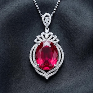 May include: A silver necklace with a large, oval, ruby-red gemstone pendant. The pendant is surrounded by a halo of small, sparkling white gemstones. The necklace is displayed against a dark background.