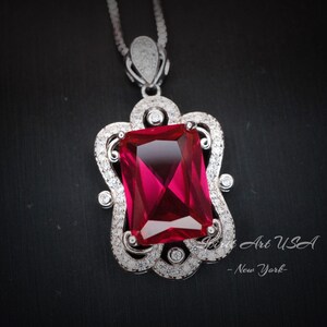 May include: A silver pendant necklace with a large, red, rectangular gemstone set in a silver filigree frame. The pendant is surrounded by smaller, clear gemstones.