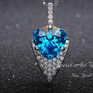May include: A silver pendant necklace with a large blue gemstone in the shape of a heart. The gemstone is surrounded by smaller clear gemstones. The pendant is in the shape of an arrowhead.