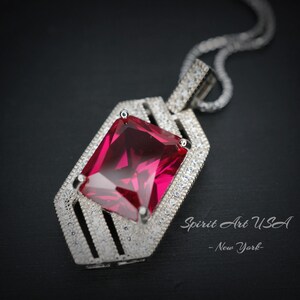 May include: A silver pendant necklace with a large, red, square-shaped gemstone set in a geometric frame. The pendant is surrounded by smaller, clear gemstones. The necklace is on a black background. Spirit Art USA - New York - is printed below the pendant.