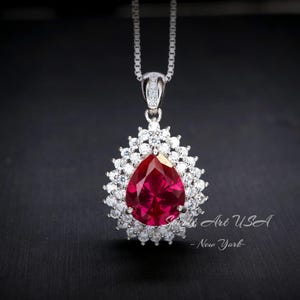 May include: A silver necklace with a teardrop-shaped red gemstone pendant surrounded by smaller clear gemstones.