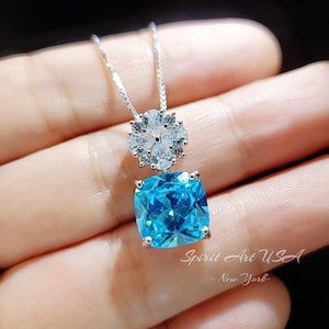 May include: A silver necklace with a blue gemstone pendant and a cluster of clear cubic zirconia stones. The pendant is a square-shaped cushion cut gemstone with a silver setting. The necklace is on a silver chain.