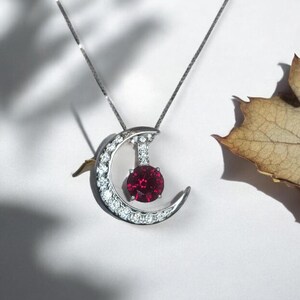 May include: A silver crescent moon pendant necklace with a red gemstone in the center and a row of clear gemstones around the edge.
