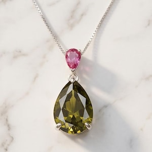 May include: A silver necklace featuring a large, olive-green pear-shaped gemstone pendant. Above it, a smaller, pink teardrop-shaped stone is suspended. The necklace is displayed on a white marble surface.