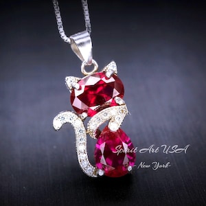 May include: A silver necklace with a red gemstone cat pendant. The cat is sitting with its tail curled around its body. The pendant is surrounded by smaller clear gemstones.