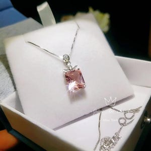 May include: A silver necklace with a pink square-shaped gemstone pendant. The pendant is surrounded by smaller clear gemstones. The necklace is in a white box with the text "Spring Art & Craft - New York" on the bottom.