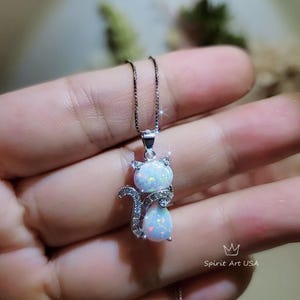 May include: A silver necklace with a white opal cat pendant. The cat is sitting with its tail curled around its body. The pendant is set with small clear stones.