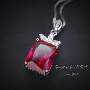 May include: A silver necklace with a red gemstone pendant. The pendant is a square cut with a silver crown design. The necklace is on a black background. Spirit Art USA - New York.