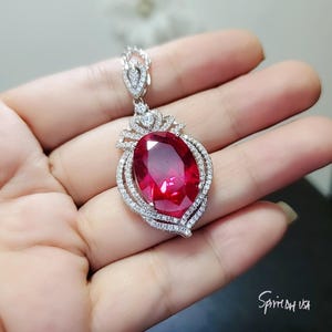 May include: A silver pendant necklace featuring a large, oval-shaped, red gemstone surrounded by smaller, clear gemstones. The pendant is set in a delicate, ornate design.