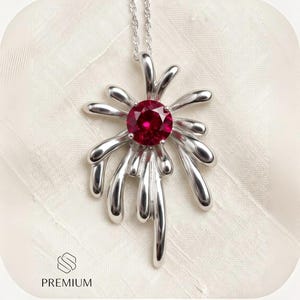 May include: A silver pendant necklace featuring a round, faceted deep red gemstone set in the center of a radiating, abstract metallic design with smooth, elongated, and curved silver prongs extending outward from the central stone.