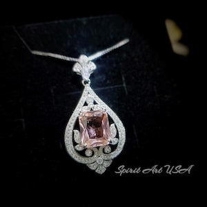 May include: A silver pendant necklace with a pink gemstone set in a square shape. The pendant is surrounded by smaller clear gemstones and has a delicate, ornate design. The necklace is on a black background with the text "Spirit Art USA" visible in the bottom right corner.