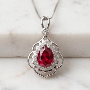 May include: A silver pendant necklace with a large, teardrop-shaped, deep red gemstone. The gemstone is encircled by small, clear stones and set in an ornate silver frame. The necklace is suspended from a fine silver chain.