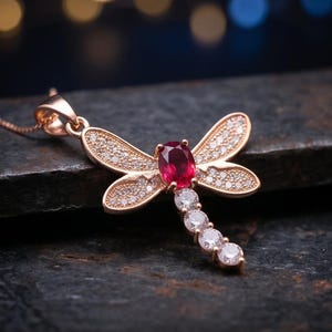 May include: A dragonfly pendant crafted in rose-toned metal featuring a central oval-cut deep red gemstone on the thorax. The four wings are encrusted with small clear pavé stones, and the tail consists of four round-cut clear stones in a row.