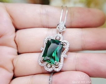 May include: A silver necklace with a large emerald green gemstone pendant set in a silver filigree frame. The pendant is surrounded by smaller clear gemstones.