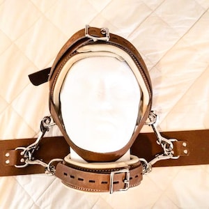 Puede incluir: Un collar de restricción de cuero marrón con un forro acolchado y una hebilla de metal. El collar está unido a dos correas de cuero marrón con hebillas de metal. Las correas están conectadas a una cadena de metal.