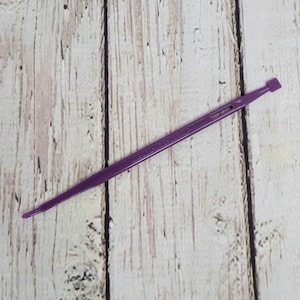 Puede incluir: Una fina herramienta de plástico morado con un extremo plano y rectangular. La herramienta está diseñada con una punta afilada en un extremo y una pequeña forma plana y rectangular en el otro. La herramienta se encuentra sobre un fondo de madera clara y envejecida.