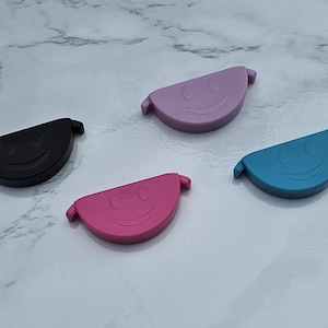 Pode incluir: Quatro rolhas de garrafa de silicone semicirculares coloridas. As rolhas são pretas, rosa, roxas e azuis, cada uma com um design de rosto sorridente. Elas têm pequenas alças em cada lado. As rolhas estão em uma superfície de mármore branco e cinza.